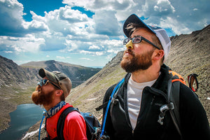 Two Men hiking in the mountains with a clear blue sky, while wearing colored sunscreen on their noses. Eco friendly sunscreen, Best SPF for face, healthy sunscreen.